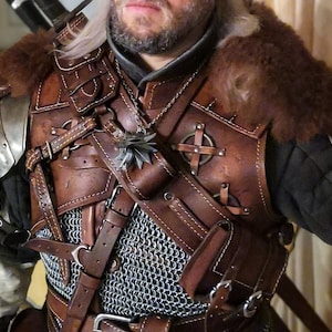 May include: A brown leather armor with metal accents and a chainmail shirt underneath. The armor has straps and buckles, and a pouch with a wolf head symbol is attached to the shoulder strap.