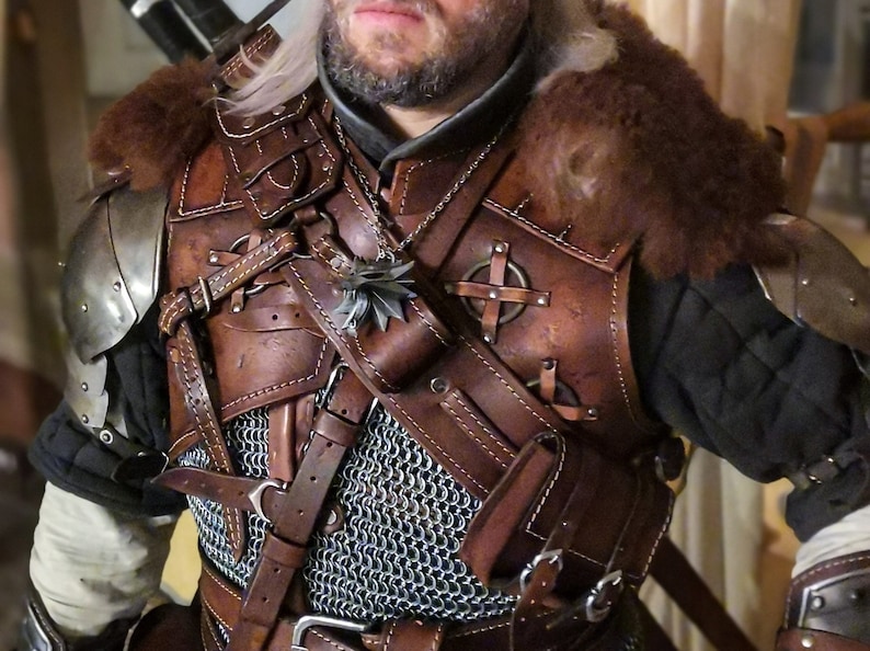 May include: A brown leather armor with metal accents and a chainmail shirt underneath. The armor has straps and buckles, and a pouch with a wolf head symbol is attached to the shoulder strap.