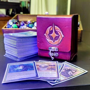 May include: A burgundy leather deck box with a gold emblem and a silver clasp. The box is sitting on a table with a stack of trading cards, including "Spada dell'Animista" and "Sguardo del Mirari".