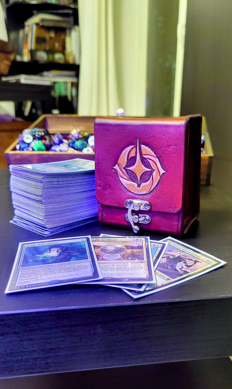 May include: A burgundy leather deck box with a gold emblem and a silver clasp. The box is sitting on a table with a stack of trading cards, including "Spada dell'Animista" and "Sguardo del Mirari".