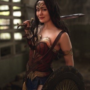 Op de afbeelding: Een vrouw verkleed als Wonder Woman, met een zwaard en schild. Ze draagt een rood, goud en blauw kostuum met een stervormige tiara. De achtergrond is een donkere, industri&euml;le omgeving.