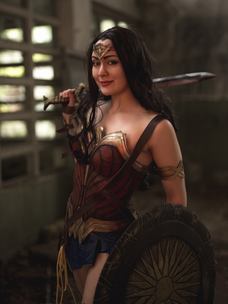 Op de afbeelding: Een vrouw verkleed als Wonder Woman, met een zwaard en schild. Ze draagt een rood, goud en blauw kostuum met een stervormige tiara. De achtergrond is een donkere, industri&euml;le omgeving.