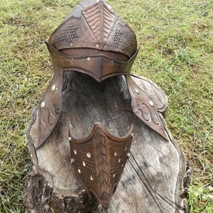 May include: A brown leather helmet with a textured design and metal accents. The helmet is on a wooden stump. There is a matching face mask next to the helmet.