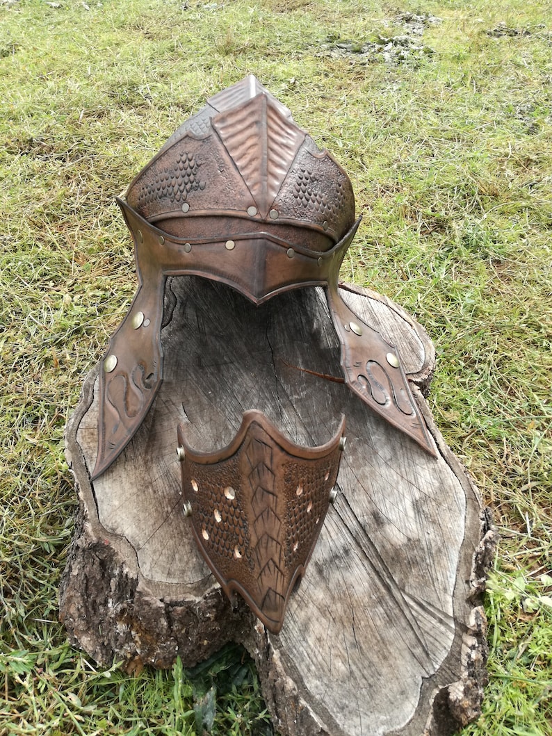 May include: A brown leather helmet with a textured design and metal accents. The helmet is on a wooden stump. There is a matching face mask next to the helmet.