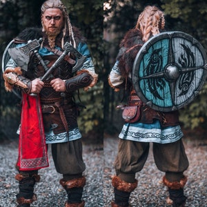 May include: A person dressed as a Viking warrior with a braided blonde wig, holding two axes and wearing a brown leather belt with a red scarf. The person is also wearing a blue and white tunic with fur trim and brown leather boots. The person is standing in front of a green background.