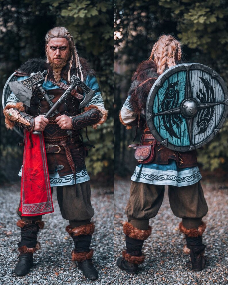 May include: A person dressed as a Viking warrior with a braided blonde wig, holding two axes and wearing a brown leather belt with a red scarf. The person is also wearing a blue and white tunic with fur trim and brown leather boots. The person is standing in front of a green background.