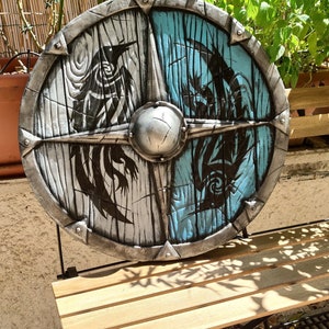 May include: A round, wooden shield with a silver metal rim. The shield is painted with a black and white design on one side and a blue and black design on the other side.