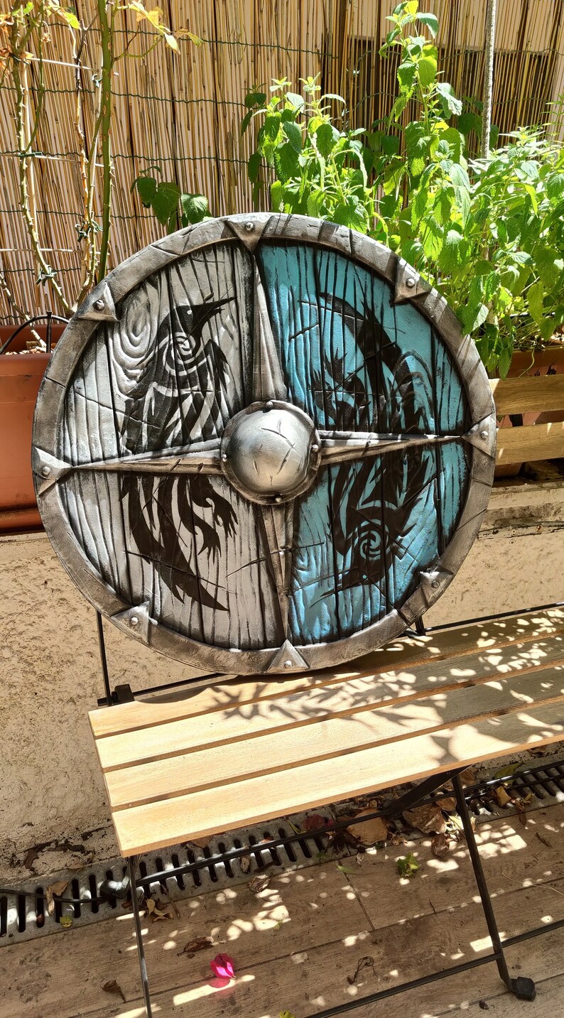 May include: A round, wooden shield with a silver metal rim. The shield is painted with a black and white design on one side and a blue and black design on the other side.