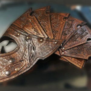 May include: A brown leather helmet with intricate designs and metal accents. The helmet has a face mask and a series of overlapping plates on the back.