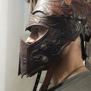 May include: A brown leather helmet with intricate carvings and metal accents. The helmet has a face mask with a pointed chin guard and a strap that goes under the chin.
