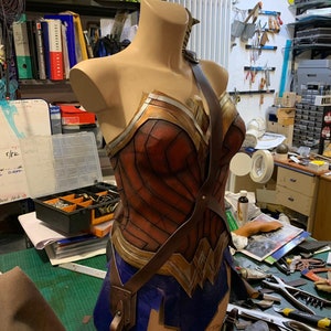 Op de afbeelding: Een bruin leren Wonder Woman-kostuum met een gouden en rode buste en een blauwe rok. Het kostuum is op een mannequin in een werkplaats.
