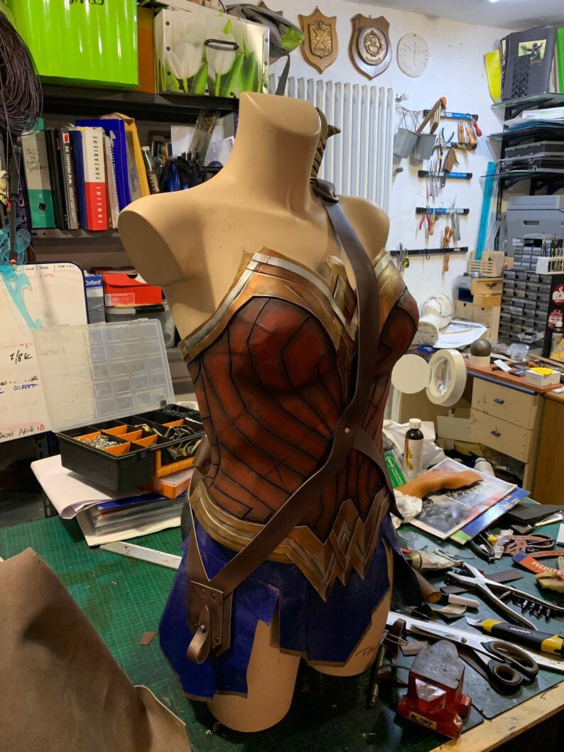 Op de afbeelding: Een bruin leren Wonder Woman-kostuum met een gouden en rode buste en een blauwe rok. Het kostuum is op een mannequin in een werkplaats.