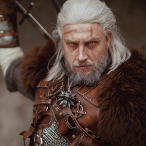 May include: A man with long white hair and a beard is wearing a brown leather and fur costume. He has a sword strapped to his back and a wolf pendant around his neck. He is looking directly at the camera with a serious expression.