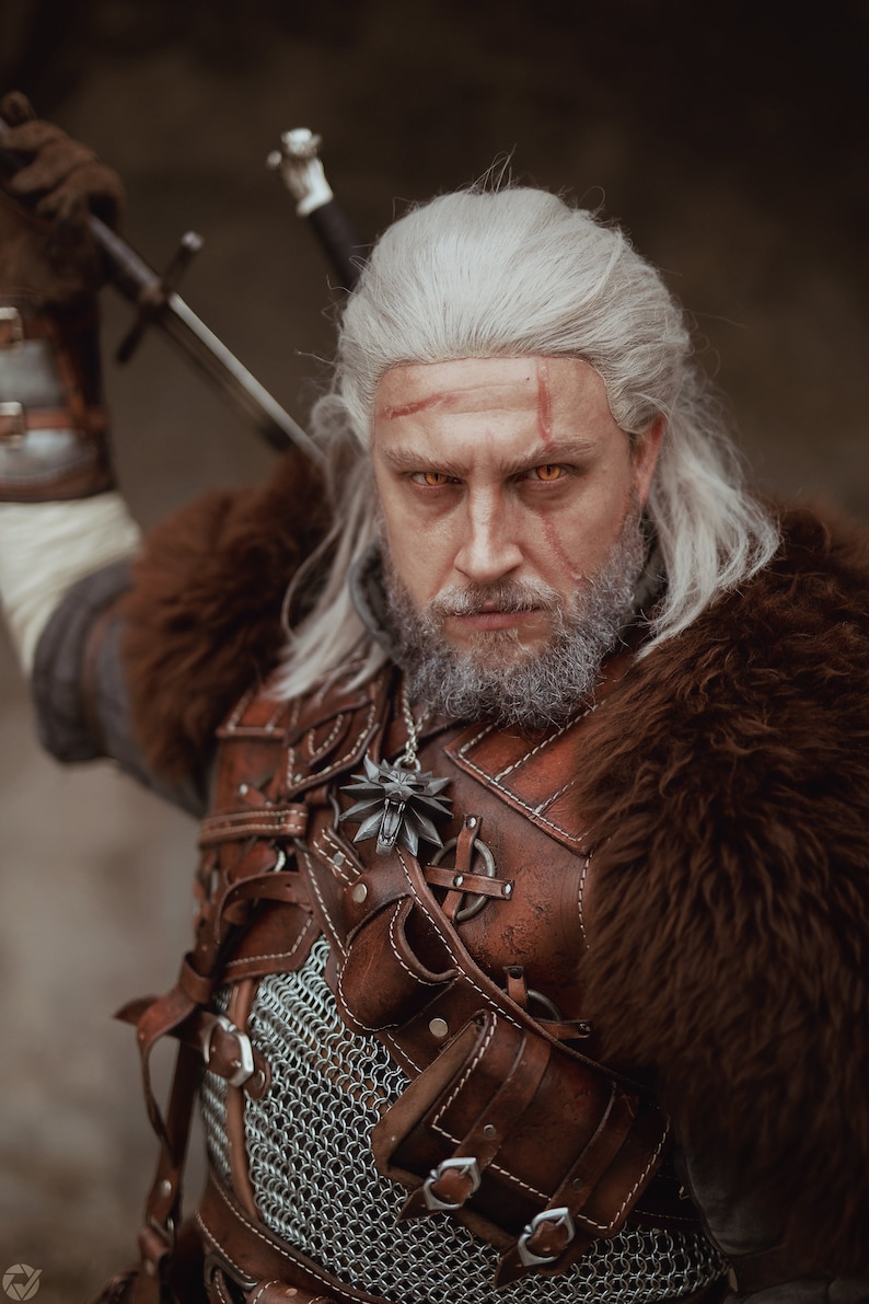 May include: A man with long white hair and a beard is wearing a brown leather and fur costume. He has a sword strapped to his back and a wolf pendant around his neck. He is looking directly at the camera with a serious expression.