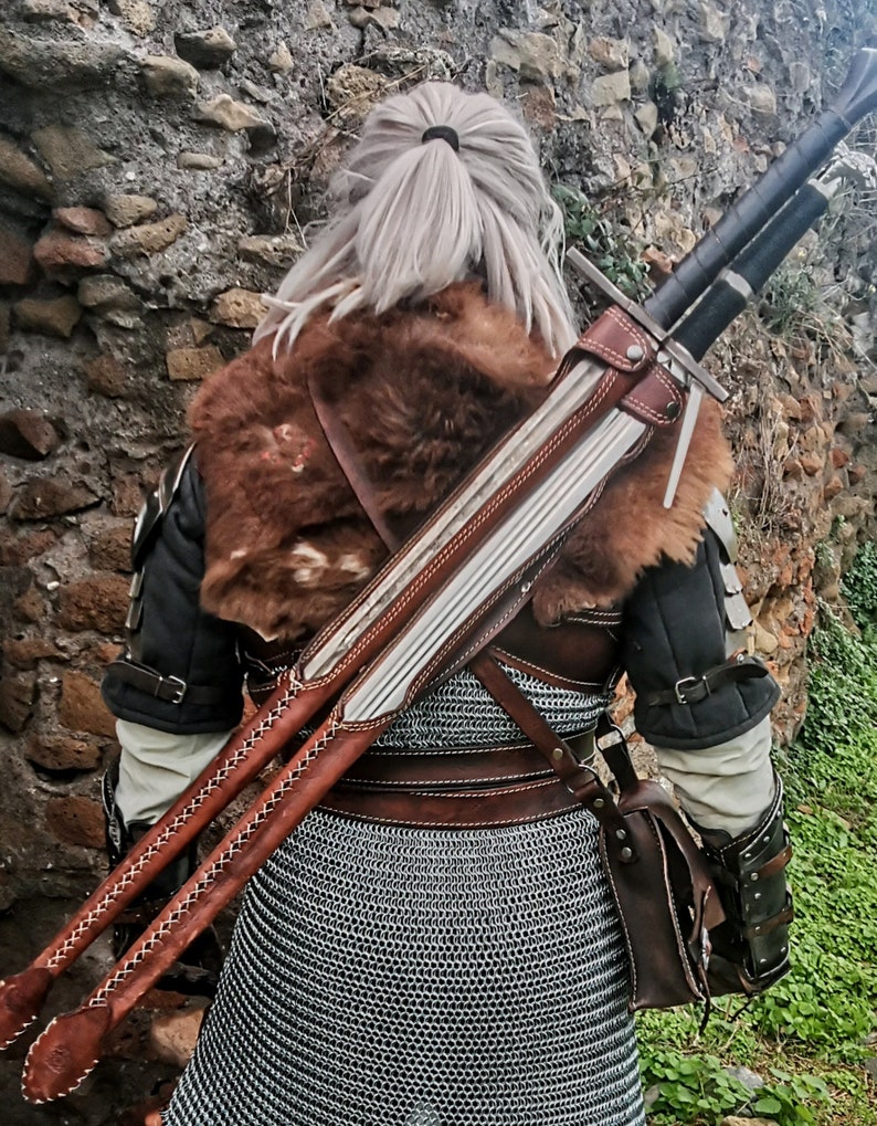 May include: A brown leather scabbard with two silver swords strapped to the back of a person wearing a brown fur-lined jacket and chainmail.