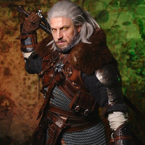 May include: A man dressed as a fantasy character, Geralt of Rivia, from the Witcher series. He is wearing a brown leather jerkin with chainmail armor underneath, a fur-lined collar, and a sword strapped to his back. He has white hair and a beard, and his eyes are a bright yellow.