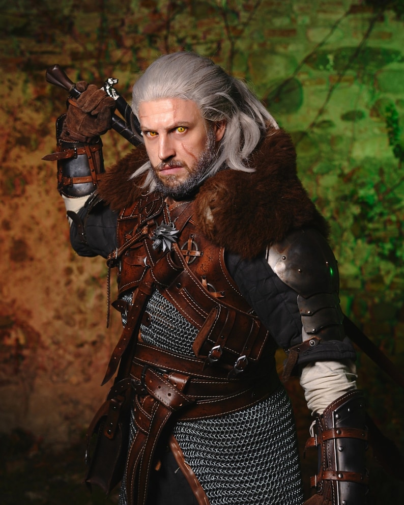 May include: A man dressed as a fantasy character, Geralt of Rivia, from the Witcher series. He is wearing a brown leather jerkin with chainmail armor underneath, a fur-lined collar, and a sword strapped to his back. He has white hair and a beard, and his eyes are a bright yellow.