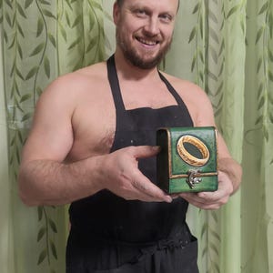 May include: A person wearing a black apron holds a small green box with a gold ring and inscription on the lid. The ring is a replica of the One Ring from the Lord of the Rings.