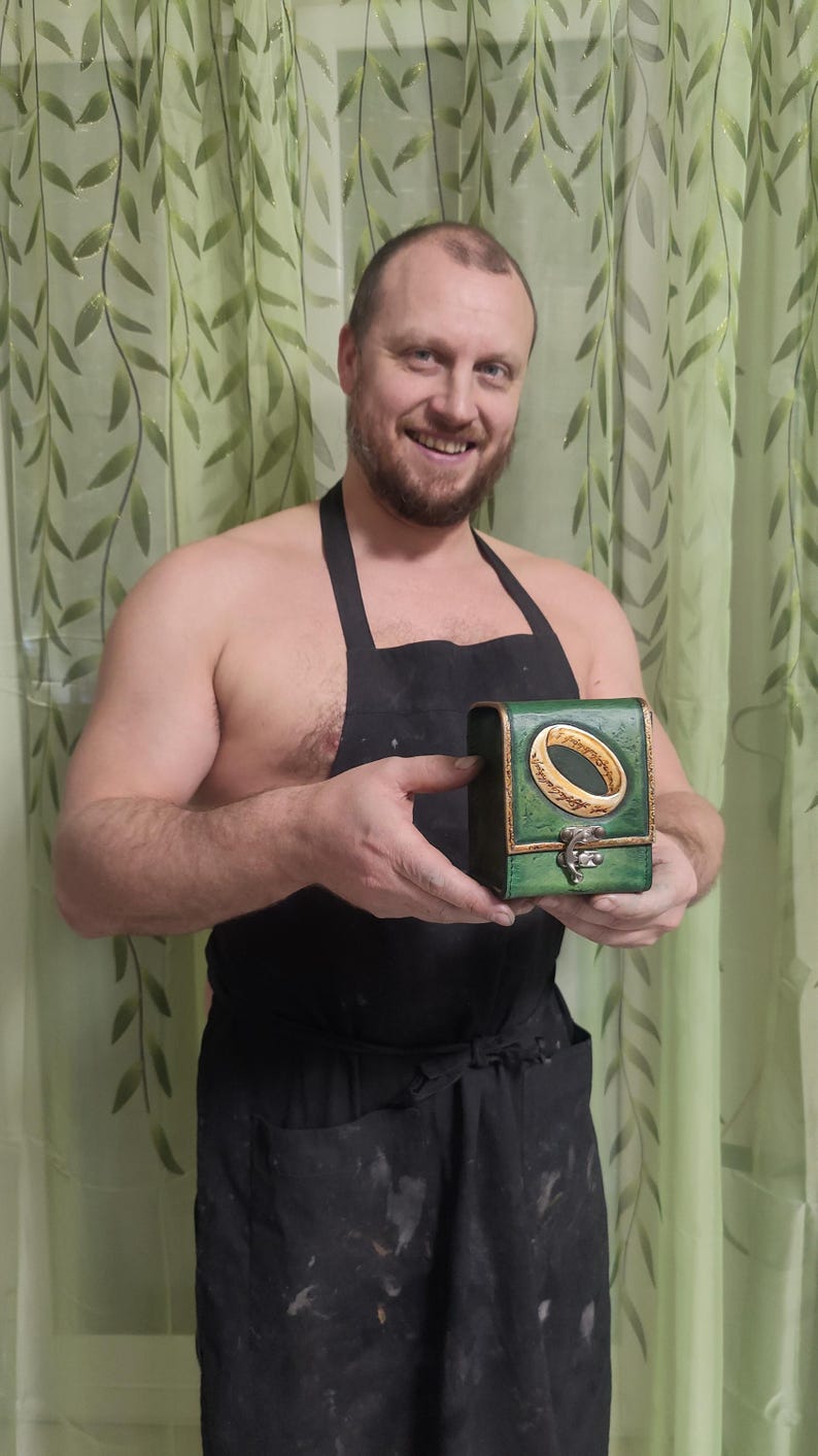 May include: A person wearing a black apron holds a small green box with a gold ring and inscription on the lid. The ring is a replica of the One Ring from the Lord of the Rings.