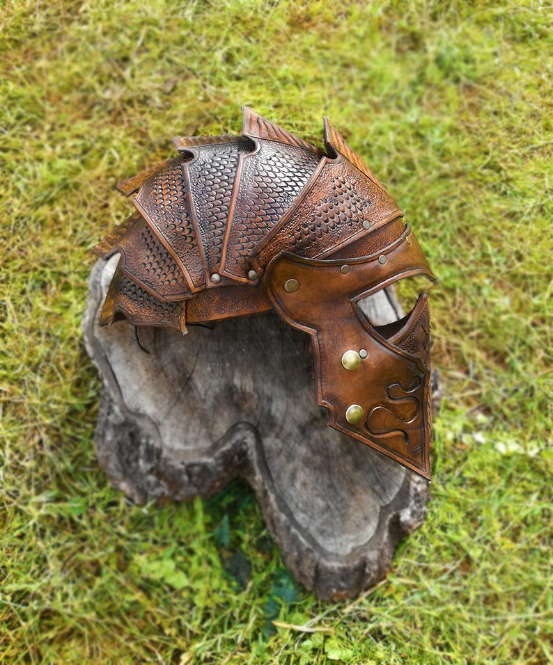 May include: A brown leather helmet with a dragon scale design. The helmet has a face mask with a decorative pattern and brass accents.