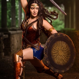 Op de afbeelding: Een vrouw verkleed als Wonder Woman, met een zwaard in de ene hand en een schild in de andere. Ze draagt een rood, goud en blauw kostuum met een gouden tiara. Ze staat in een bosrijke omgeving.