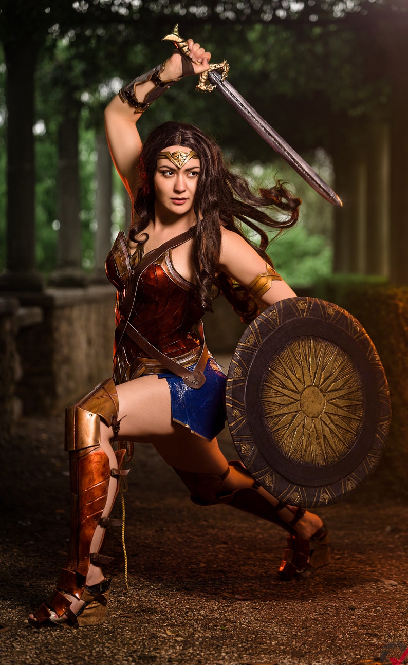 Op de afbeelding: Een vrouw verkleed als Wonder Woman, met een zwaard in de ene hand en een schild in de andere. Ze draagt een rood, goud en blauw kostuum met een gouden tiara. Ze staat in een bosrijke omgeving.