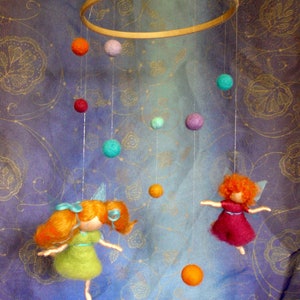Op de afbeelding: Een kleurrijk vilt mobiel met twee vilt feeën, een in een groene jurk en een in een rode jurk, hangend aan een houten ring. Het mobiel heeft ook vilt ballen in verschillende kleuren, waaronder oranje, rood, paars, groen en blauw.