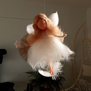 Peut inclure: Une poupée de fée en feutre blanc avec des cheveux blonds et une robe blanche, suspendue au plafond. La fée est debout sur un croissant de lune avec une étoile au-dessus de sa tête.