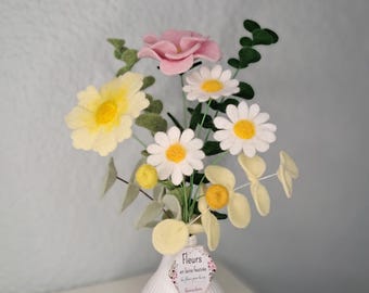 May include: A white vase with a bouquet of felt flowers. The bouquet includes yellow daisies, white daisies with yellow centers, a pink rose, and green leaves. A tag on the vase reads "Fleurs en laine feutrée".