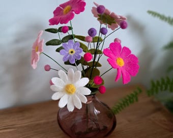May include: A small, brown glass vase holding a colorful arrangement of felt flowers. The flowers include pink, purple, and white blooms with yellow centers, and green leaves. The vase sits on a wooden surface.