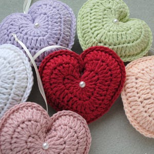 May include: Six crocheted heart-shaped ornaments in various colours, including white, lavender, green, red, and pink. Each heart has a pearl button in the centre.