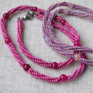 Puede incluir: Dos hileras de collares de cuentas rosadas y moradas con acentos de piedras preciosas redondas.