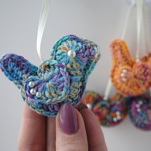 May include: Five crocheted bird ornaments with pearl accents. The birds are in shades of blue, purple, orange, and pink. They are hanging from white ribbon.