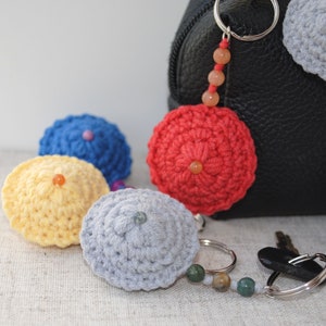 May include: A set of crocheted keyrings in various colours, including red, yellow, blue, and grey. The keyrings are shaped like small hats and feature a beaded accent. The keyrings are attached to a silver keyring with a beaded charm.