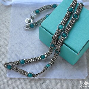 May include: A silver necklace with turquoise blue beads and a silver clasp. The necklace is displayed on a turquoise blue box and a white fabric background.