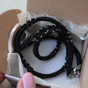 May include: A black beaded necklace with silver accents and a decorative clasp, presented in a small, open cardboard box lined with white fabric. The necklace is coiled within the box, ready for gifting.