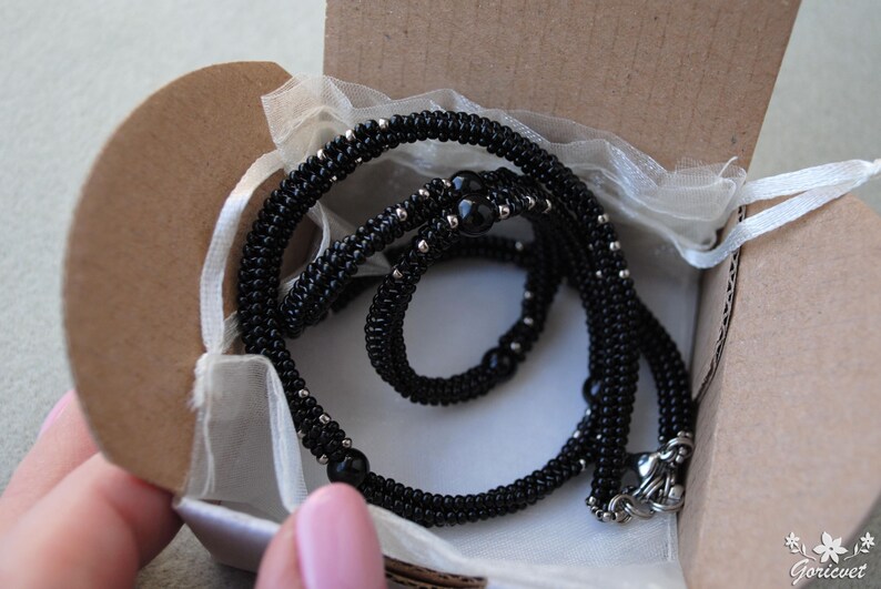 May include: A black beaded necklace with silver accents and a decorative clasp, presented in a small, open cardboard box lined with white fabric. The necklace is coiled within the box, ready for gifting.