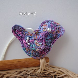 May include: A crocheted butterfly brooch in shades of purple, pink, and blue with white pearl accents. The brooch is attached to a wooden and wicker base.