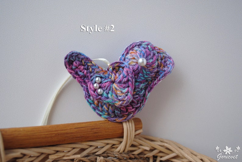 May include: A crocheted butterfly brooch in shades of purple, pink, and blue with white pearl accents. The brooch is attached to a wooden and wicker base.