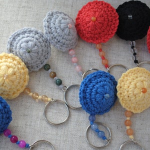 May include: A collection of nine crocheted keyrings in various colours, including blue, yellow, red, grey, and black. Each keyring features a small crocheted ball with a bead in the centre and is attached to a metal ring with a beaded chain.
