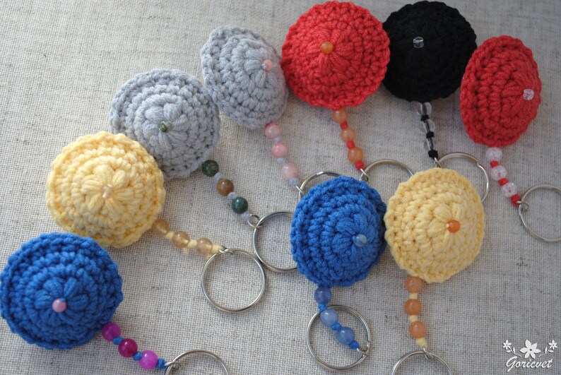 May include: A collection of nine crocheted keyrings in various colours, including blue, yellow, red, grey, and black. Each keyring features a small crocheted ball with a bead in the centre and is attached to a metal ring with a beaded chain.