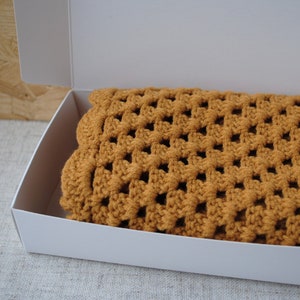 May include: A crocheted blanket in a warm brown color is folded and placed inside a white box. The blanket has a square pattern.