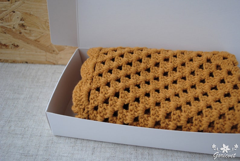 May include: A crocheted blanket in a warm brown color is folded and placed inside a white box. The blanket has a square pattern.