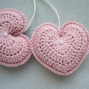May include: Two pink crocheted hearts with white pearl embellishments. The hearts are hanging from white string.