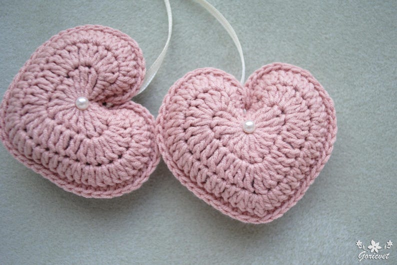 May include: Two pink crocheted hearts with white pearl embellishments. The hearts are hanging from white string.