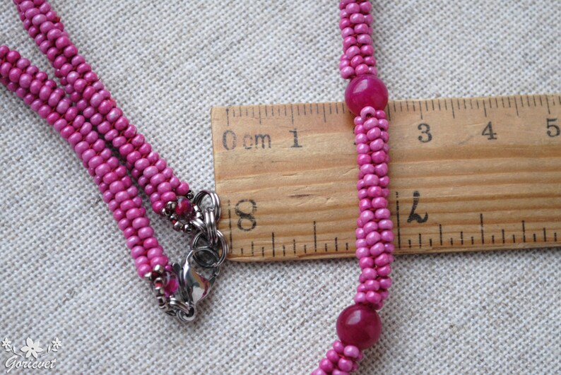 Puede incluir: Un collar de cuentas rosadas con dos cuentas rosadas m&aacute;s grandes y un cierre plateado. El collar se mide con una regla, mostrando que mide aproximadamente 5 cm de largo.