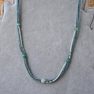 May include: A delicate necklace made with small, light blue and white beads. The necklace has four larger, round, green beads spaced evenly along the length.