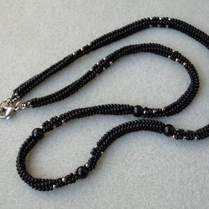 May include: A black beaded necklace with silver accents and a silver clasp. The necklace features a woven design with small black beads and larger black beads spaced throughout. The jewelry is displayed on a light gray surface.
