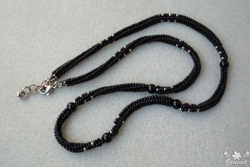 May include: A black beaded necklace with silver accents and a silver clasp. The necklace features a woven design with small black beads and larger black beads spaced throughout. The jewelry is displayed on a light gray surface.
