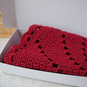 Pu&ograve; includere: Una coperta di uncinetto rossa piegata in una scatola bianca. La coperta ha un motivo a pizzo.